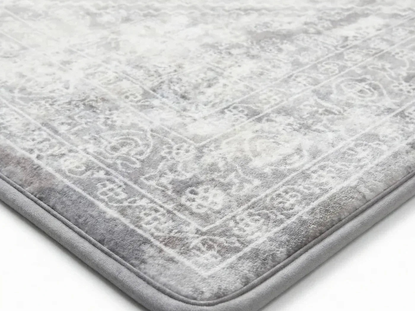 Close-up of a CloudMat corner on a pale floor: a single bonded piece with a soft patterned microfibre top and a narrow bound edge running around it, with no seam anywhere between a cover and a pad
