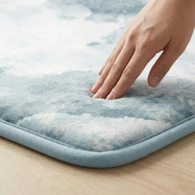 A hand pressing into the soft patterned surface of a CloudMat, the fingers sinking into the pile, with the bound edge and the full thickness of the rug visible in profile against a wood floor